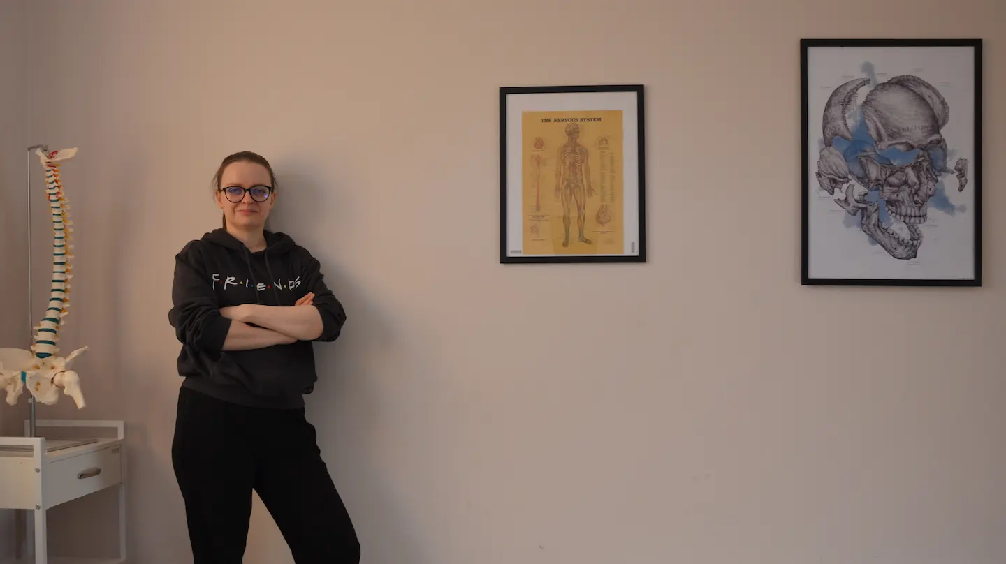 Zdjęcie Anny Czajkowskiej przy stole do masażu z atrybutami: modelem kręgosłupa, obrazami z modelmi anatomicznymi